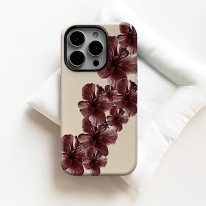 Puede incluir: Funda de teléfono beige con flores de hibisco granate. La funda tiene un recorte para la cámara con tres lentes y un flash. El diseño floral está en la parte posterior de la funda.