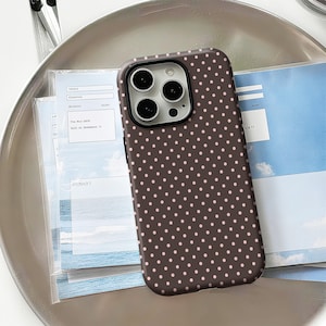 May include: A brown phone case with a pattern of small, evenly spaced pink polka dots. The case has a black border around the camera lenses. The phone case is resting on a silver tray with some papers.