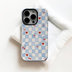 Op de afbeelding: Een telefoonhoesje met een blauw en wit geruit patroon. De hoes heeft rode harten en kersen vastgebonden met blauwe strikken. De cameralens is zichtbaar aan de bovenkant. De hoes ligt op een wit oppervlak.