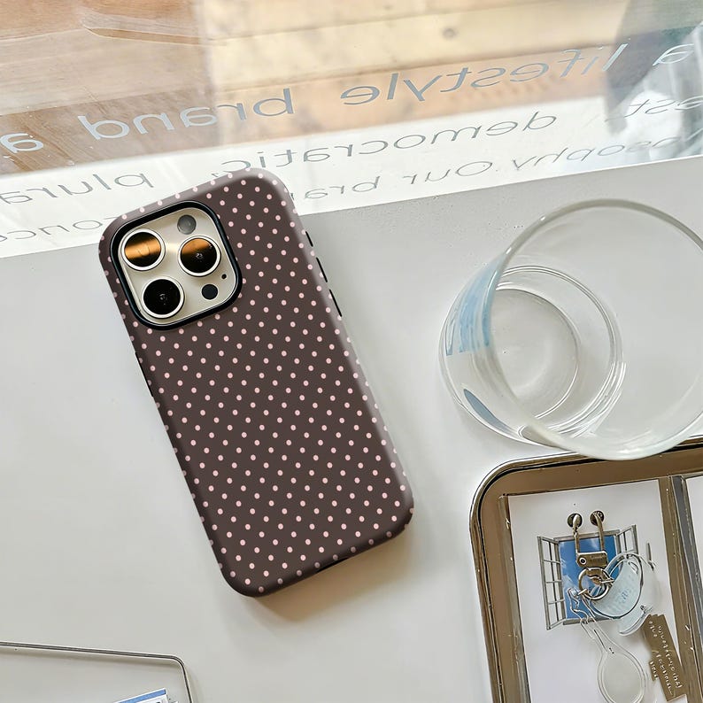 May include: A brown phone case with a polka dot pattern. The case has a dark border and a camera cutout. The phone case is lying on a white surface.