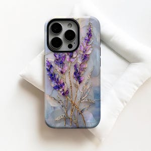 Può includere: Una custodia per telefono con un design floreale con fiori di lavanda viola e dettagli dorati. La custodia ha uno sfondo effetto marmo e una cornice nera per l'obiettivo della fotocamera. La custodia è su una superficie bianca.