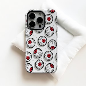 May include: A smartphone case featuring a repeating pattern of white eyeballs with red irises and black pupils on a light grey background. The case has a black camera cutout area at the top left, designed to fit a device with a triple-lens camera system.