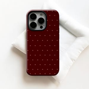 May include: A burgundy phone case with a repeating pattern of small, light-colored dots. The case has a glossy finish and a camera cutout with multiple lenses. The phone case is resting on a white surface.