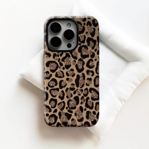 Peut inclure: Coque de téléphone avec un motif léopard. L'étui présente un fond beige avec des taches noires et dorées scintillantes. L'étui est posé sur une surface blanche.