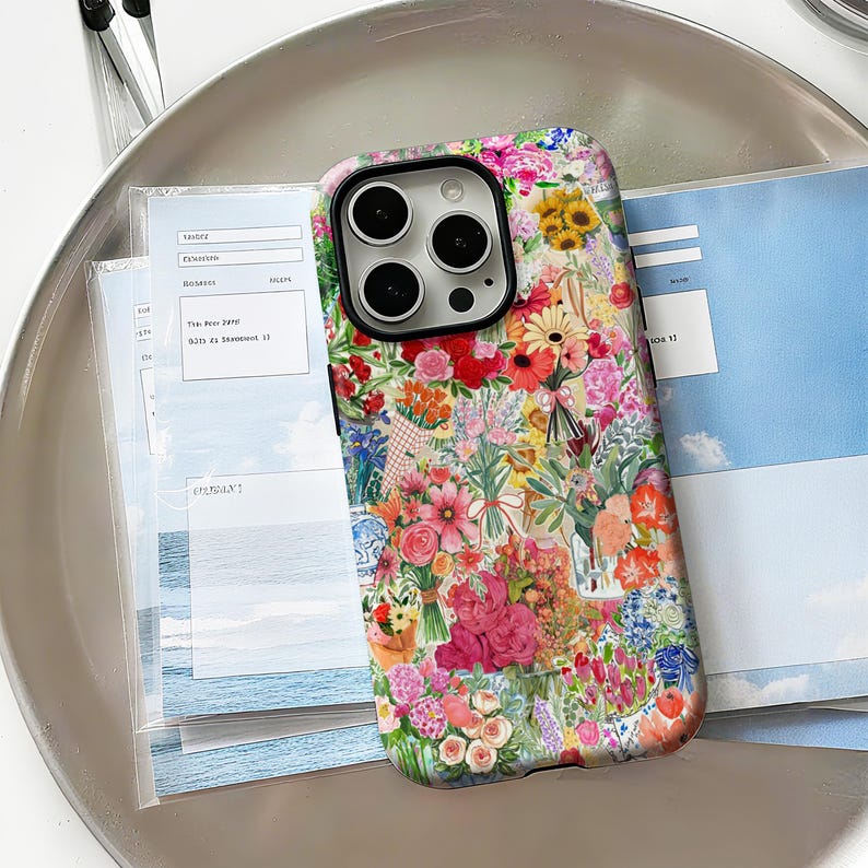 May include: A floral phone case featuring a vibrant collage of flowers in various colors, including pink, red, yellow, and blue. The case has a black camera module and is set against a light background with some papers.