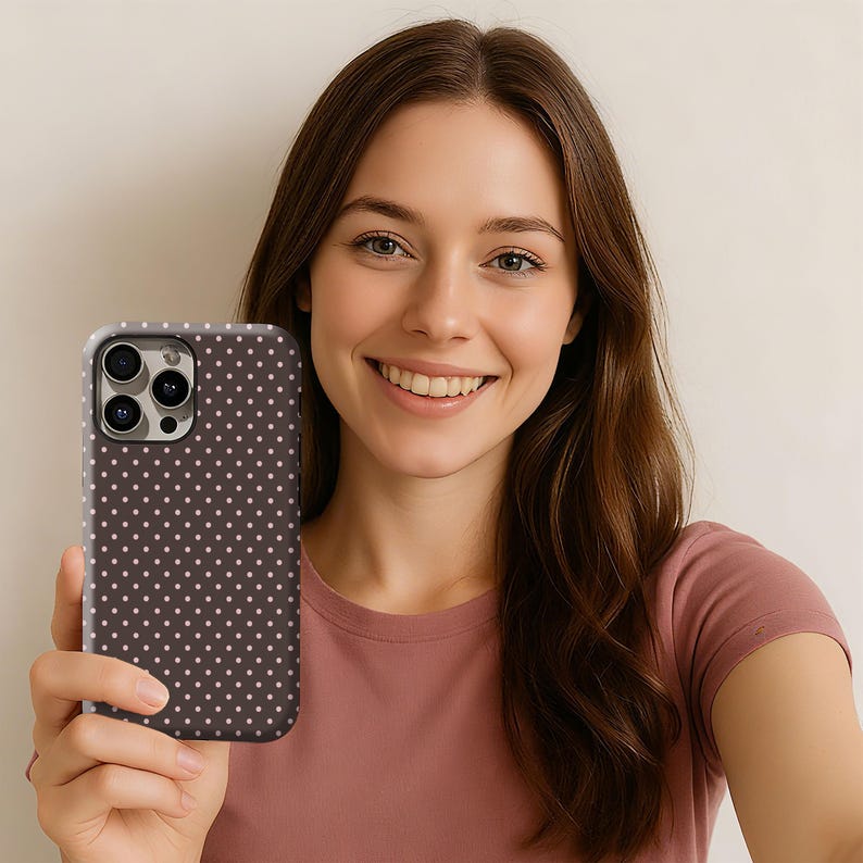 May include: A brown phone case with a white polka dot pattern. The case is held by a person wearing a pink shirt. The phone case has a camera cutout and a protective bumper.