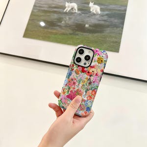 May include: A floral phone case featuring a vibrant array of pink, red, yellow, and blue flowers. The case has a black camera module and is held in a person's hand. The background includes a framed picture with two white rabbits.