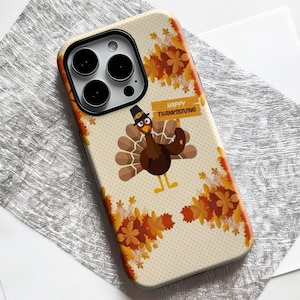 May include: A phone case featuring a Thanksgiving theme. The case has a beige background with a cartoon turkey wearing a pilgrim hat and holding a sign that says "Happy Thanksgiving." The design includes fall leaves in shades of orange and brown.