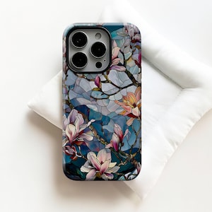 Può includere: Custodia per smartphone con un design a vetrata raffigurante fiori di magnolia rosa e gialli su sfondo blu. La custodia ha una finitura lucida e un bordo scuro, con le lenti della fotocamera visibili.
