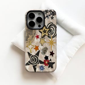 Puede incluir: Funda de teléfono con fondo crema con un collage de elementos celestiales y disco. Estrellas doradas, naranjas y negras, una luna y una pequeña bola de discoteca. La funda tiene un módulo de cámara negro.