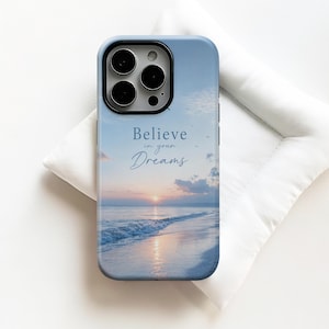 Może przedstawiać: Etui na smartfona z nadrukiem plaży o zachodzie słońca w delikatnych odcieniach błękitu i różu. Napis Believe in your Dreams jest wydrukowany ciemnoniebieską czcionką szeryfową i kursywą na środku powierzchni etui ochronnego.