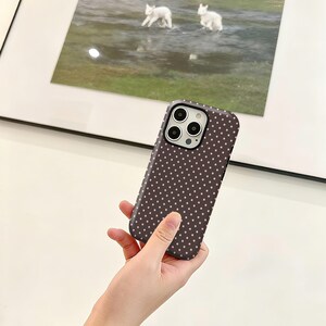 May include: A dark brown phone case with a pattern of small, evenly spaced white dots. The case is held in a hand, and the camera lenses are visible. The background includes a framed picture with a landscape scene.