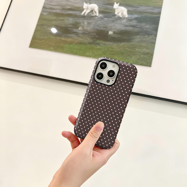 May include: A dark brown phone case with a pattern of small, evenly spaced white dots. The case is held in a hand, and the camera lenses are visible. The background includes a framed picture with a landscape scene.