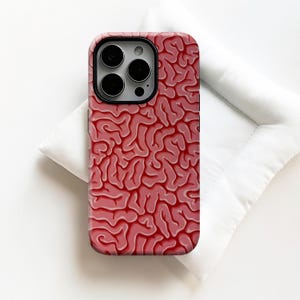 May include: A smartphone case with a brain pattern design in shades of red and pink. The case has a glossy finish and is shown on a white surface. The design mimics the folds and contours of a brain.