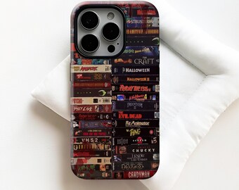 Puede incluir: Una funda de teléfono con un collage de cintas VHS vintage, incluyendo títulos como "Halloween", "Viernes 13" y "Candyman". La funda tiene un acabado brillante y un fondo oscuro y texturizado, evocando un tema de película de terror retro.