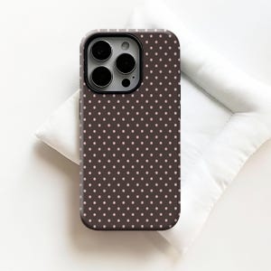 May include: A brown phone case with a repeating pattern of small, light pink polka dots. The case has a black camera lens frame and is designed to fit a smartphone. The case is shown on a white surface.