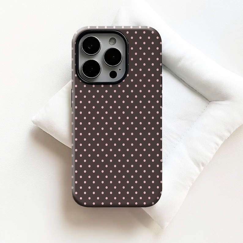 May include: A brown phone case with a repeating pattern of small, light pink polka dots. The case has a black camera lens frame and is designed to fit a smartphone. The case is shown on a white surface.