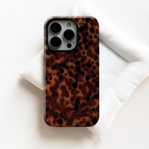 Op de afbeelding: Een telefoonhoesje met een bruin en zwart schildpadpatroon. De hoes heeft een glanzende afwerking en een donkere rand rond de cameralenzen. De telefoonhoes ligt op een witte ondergrond.