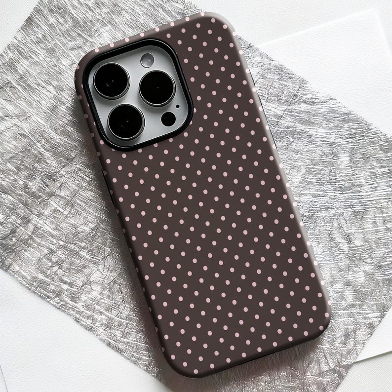 May include: A brown phone case with a pattern of small, evenly spaced pink polka dots. The case has a dark brown border around the camera and a silver camera lens. The case is lying on a textured silver surface.