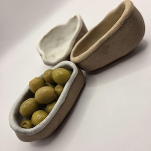 Pode incluir: Três pequenas travessas de cerâmica feitas à mão. Uma travessa é preenchida com azeitonas verdes. As travessas têm formato oval com exterior castanho claro e borda interna branca. As azeitonas são de cor verde vibrante.