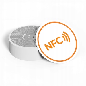 Op de afbeelding: Een stapel witte NFC-stickers met een grijs, getextureerd ontwerp op de rand. Eén sticker is los, met een oranje ring rond de witte cirkel met de oranje tekst "NFC" en signaalbalken.