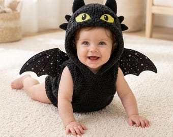May include: A black sleeveless hooded romper featuring a textured fleece material, two black horns on top, and large yellow eyes on the hood. The garment includes two attached black wings with a white star pattern on the back side of the wings.