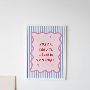 May include: A framed wall art print featuring a light blue and white vertical striped background with a central pink scalloped rectangle. The text reads "NOS DA, CARU TI, WELAI DI YN Y BORE" in red, accented by small yellow stars and pink hearts.