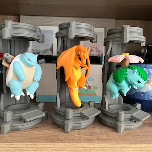 May include: Three gray 3D-printed cylindrical display pods, each containing a different character figure. The left pod holds a light blue turtle-like creature, the center pod holds an orange dragon-like creature, and the right pod holds a green creature with a pink flower on its back.