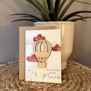 Könnte beinhalten: Eine Grußkarte mit einem Holz-Heißluftballon-Design. Die Karte hat einen cremefarbenen Hintergrund mit aquarellartigen roten Wolken und dem Text "Willkommen Kleine Maus". Die Karte ist vor einem braunen Kartonhintergrund platziert.