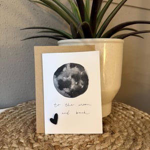 Könnte beinhalten: Eine Grußkarte mit einer Aquarellzeichnung des Mondes in Grau- und Schwarztönen. Auf der Karte steht "to the moon and back" in handschriftlicher Schrift. Ein kleines schwarzes Herz ist an der Karte befestigt. Die Karte befindet sich in einem braunen Umschlag.