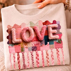 Może przedstawiać: Beżowa bluza z napisem "LOVE" wyhaftowanym różową przędzą. Litery są wypukłe i teksturowane, na tle patchworku z kolorowych kwadratów materiału. Plecione frędzle zwisają pod słowem, dodając dekoracyjnego akcentu.