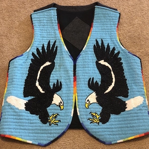 Peut inclure: Un gilet perlé bleu clair avec deux aigles noirs aux accents blancs et des serres jaunes. Le gilet a un dos noir et est bordé d'un motif arc-en-ciel. Le motif de l'aigle est centré sur les panneaux avant.
