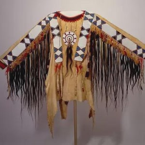 May include: A tan-colored Native American ceremonial garment with long sleeves and fringe. The sleeves are decorated with white, red, blue, and black geometric patterns. A circular pendant hangs from the neckline. The garment is displayed on a stand.