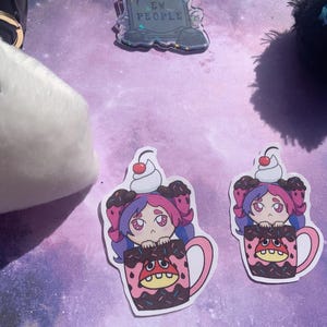 May include: A white plush ghost with black eyes and a black pom-pom. Two stickers feature a cartoon girl in a mug with whipped cream and a cherry on top. A pin with the text "EW PEOPLE" is also visible.
