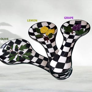 Puede incluir: Tres cucharas de servir decorativas con patrones de cuadros blancos y negros. Cada cuchara presenta un diseño de fruta diferente: aceitunas, limones y uvas. Las cucharas están dispuestas en una composición en capas.