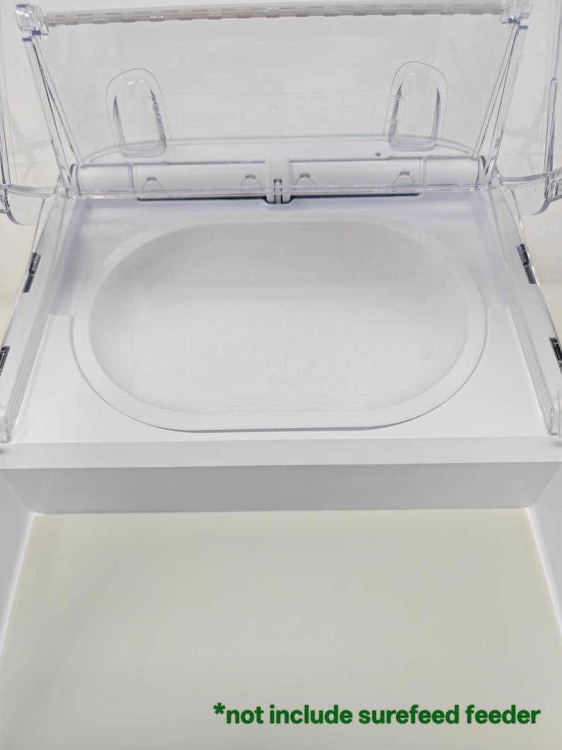 Puede incluir: Un comedero para mascotas de pl&aacute;stico transparente con interior blanco y tapa abatible. El comedero tiene un compartimento para alimentos de forma ovalada y el texto "*not include surefeed feeder" en verde.