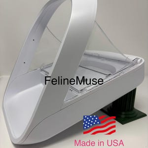 Puede incluir: Un soporte blanco para alimentador SureFeed con una cubierta de plástico transparente. El soporte tiene un asa curva y está etiquetado como "Made in USA". El soporte está diseñado para elevar un alimentador para mascotas.