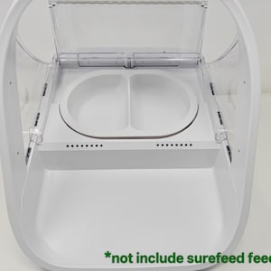 Puede incluir: Un comedero blanco para mascotas con una tapa transparente con bisagras. El comedero tiene dos compartimentos circulares para alimentos y una bandeja inferior. El texto "*not include surefeed feeder" es visible.