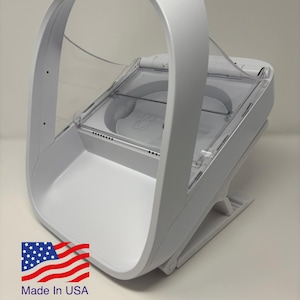 Puede incluir: Un comedero blanco para mascotas con un asa curva y una tapa transparente con bisagras. El comedero tiene una base rectangular y un diseño de huella de pata en el interior. Las palabras "Made in USA" están impresas en el lateral.