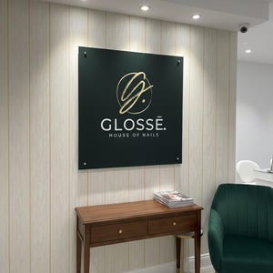 Puede incluir: Un letrero verde oscuro con letras doradas que dice "GLOSSÉ. HOUSE OF NAILS" cuelga de una pared. Debajo del letrero hay una consola de madera con dos cajones y una pila de revistas. Una silla de terciopelo verde está a la derecha.