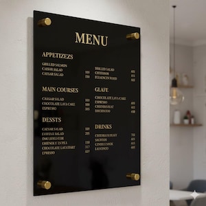 Könnte beinhalten: Eine schwarze Menütafel aus Acryl mit goldfarbenen Buchstaben und Beschlägen. Die Speisekarte listet Vorspeisen, Hauptgerichte, Desserts und Getränke auf. Die Tafel ist an einer weißen Wand befestigt.