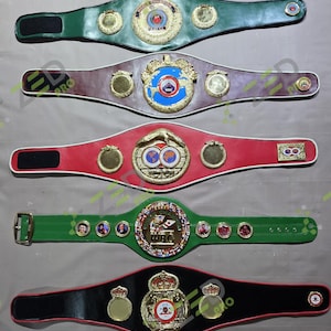 May include: Five championship belts arranged vertically. The top belt is green, followed by a brown belt, a red belt, a green belt with portrait medallions and the text WBC, and a black belt at the bottom. Each belt features gold-toned metal plates.