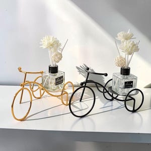 Puede incluir: Dos soportes decorativos en forma de bicicleta, uno dorado y otro negro, cada uno con una botella de vidrio transparente con detalles florales blancos. Las botellas tienen tapas negras y una etiqueta. Los soportes están sobre una superficie blanca.