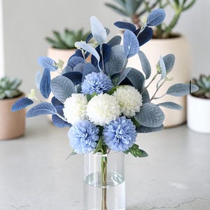 May include: A bouquet of artificial flowers in a clear glass vase filled with water. The arrangement features round white and light blue pom-pom style blossoms mixed with various shades of blue and grey-green eucalyptus leaves and stems.