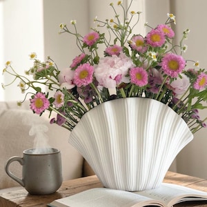 May include: A white ceramic vase with a fan-shaped, ribbed design filled with a bouquet of pink daisies, white peonies, and small white wildflowers. Beside the vase sits a grey ceramic mug with rising steam and an open book with text.