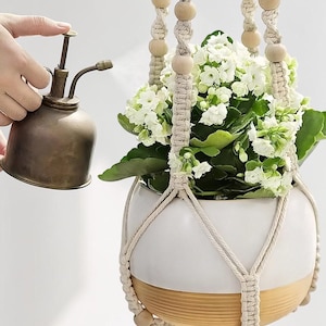 May include: A white and tan ceramic planter held by a cream-colored macrame hanger with wooden beads, containing a white flowering plant. A metallic bronze-toned spray mister is positioned to the side, emitting a fine mist toward the plant.