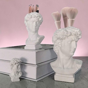 May include: Three white matte resin busts of David, each featuring a square base. The two larger busts function as holders, with one containing several wooden pencils and the other holding three makeup brushes with light-colored bristles.