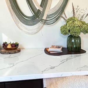 May include: A wall hanging featuring a curved dark wood frame with draped sage green and cream-colored yarn strands. The piece is displayed above a white marble countertop with a green glass vase of flowers, a wooden bowl of fruit, and books.