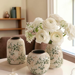 May include: Three ceramic vases with floral patterns. The vases are off-white with green and pink floral designs. One vase is cylindrical, another is round, and the third is a smaller, more rounded shape. White flowers are in one vase.