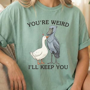 May include: Sage green t-shirt with the text "YOU'RE WEIRD I'LL KEEP YOU". The shirt features a graphic of a white duck and a gray bird with a red heart above them. The shirt has rolled-up sleeves.